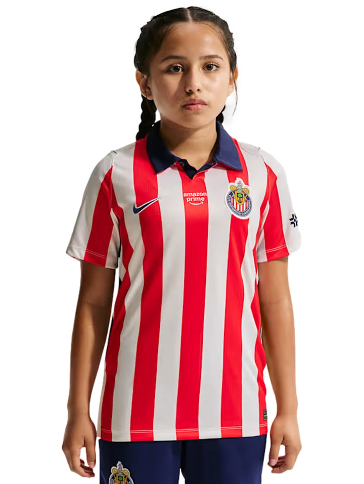 Niña vistiendo playera deportiva Nike del Chivas con rayas verticales rojas y blancas y cuello azul.