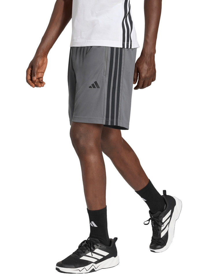 Hombre vistiendo short deportivo Adidas color gris con tres franjas negras laterales y logo frontal.